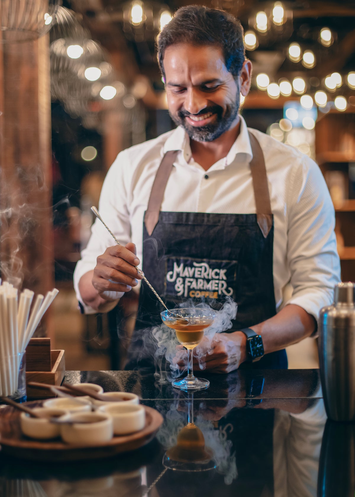 A man wearing a white shirt and a black apron with the logo 'Maverick & Farmer Coffee' smiles while preparing a drink in a bar setting with warm bokeh lights in the background.