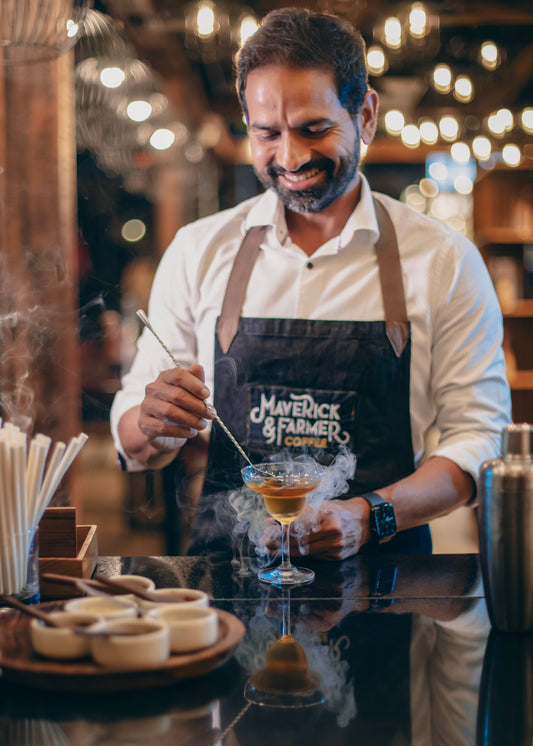 A man wearing a white shirt and a black apron with the logo 'Maverick & Farmer Coffee' smiles while preparing a drink in a bar setting with warm bokeh lights in the background.