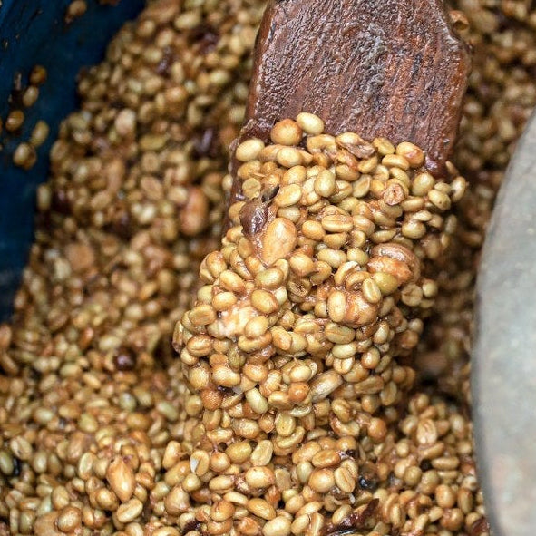 A close-up of roasted coffee beans in a blue barrel with a wooden spoon and a metal lid resting on top.
