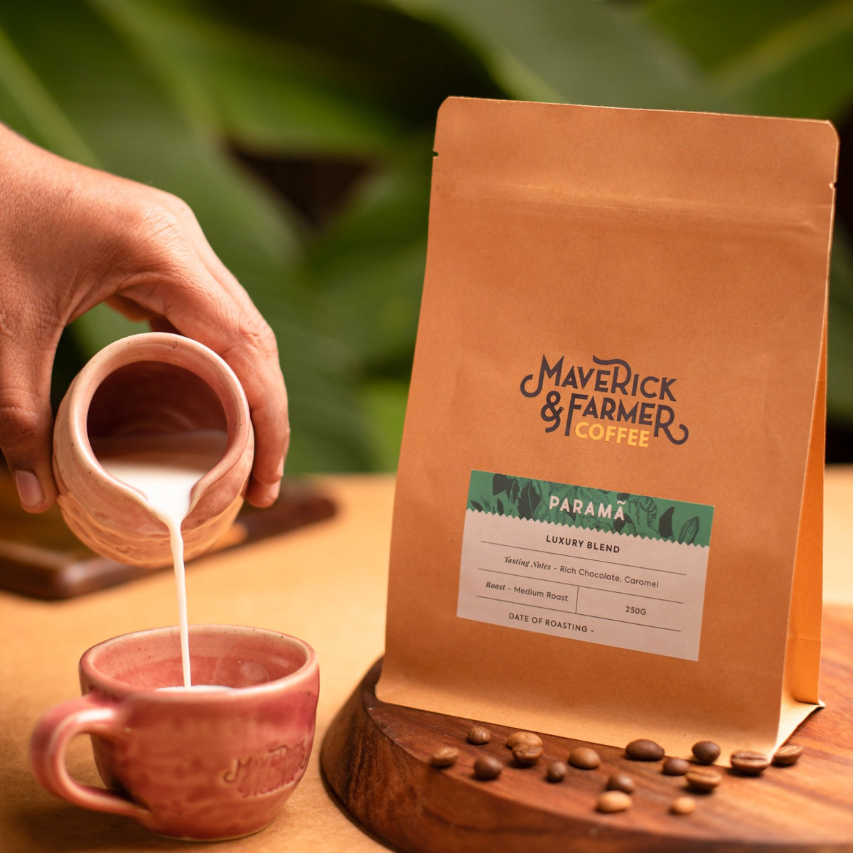 A hand pours milk from a small ceramic pitcher into a pink mug beside a standing kraft coffee bag labeled Maverick & Farmer Coffee Parama Luxury Blend, with coffee beans on a wooden surface and blurred green leaves in the background.