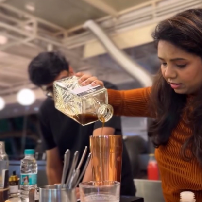 A woman in an orange ribbed sweater pours coffee from a clear glass decanter into a copper mixing cup on a counter, with bar tools and a cafe interior in the background. Text at the top reads 'Coffee Mixology Experience at @maverickandfarmer'.