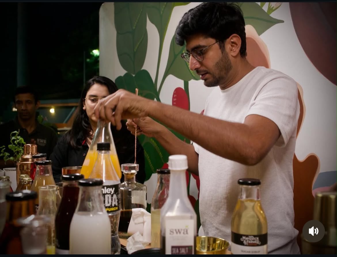 A man in a white t-shirt and glasses is pouring a yellow liquid from a glass bottle into a larger pitcher at a table filled with various bottles and glasses, with a woman and another man in the background.