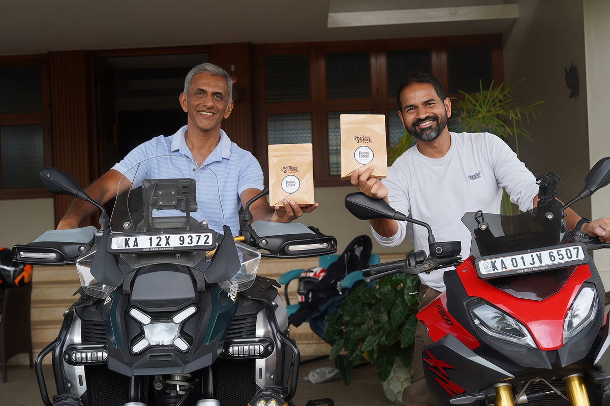 Two men with motorcycles holding a coffee bag in their hands and standing front of a house