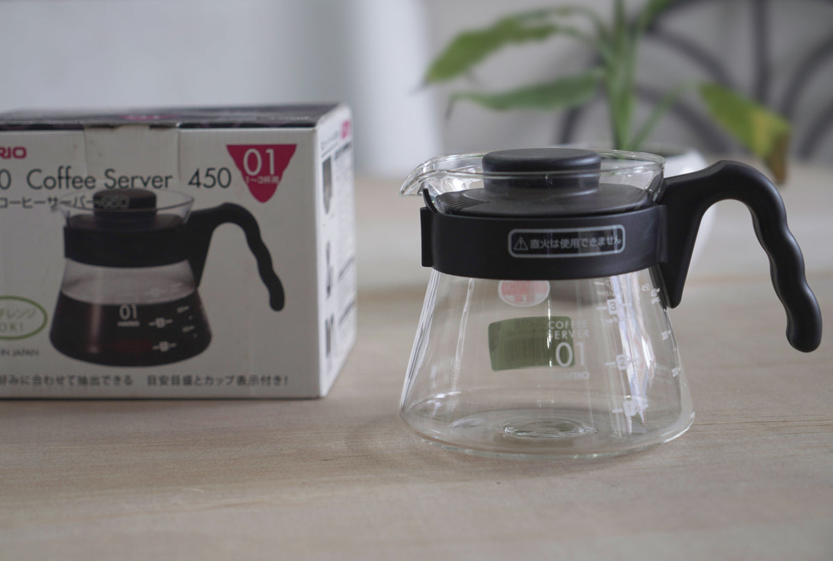 A clear glass Hario coffee server with a black plastic handle and lid, displayed alongside its packaging box.