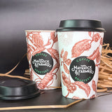 Two paper cups with floral designs and the brand 'Maverick & Farmer' coffee roasters logo, accompanied by a black sipper lid, placed on a dark surface with straw hay nearby.